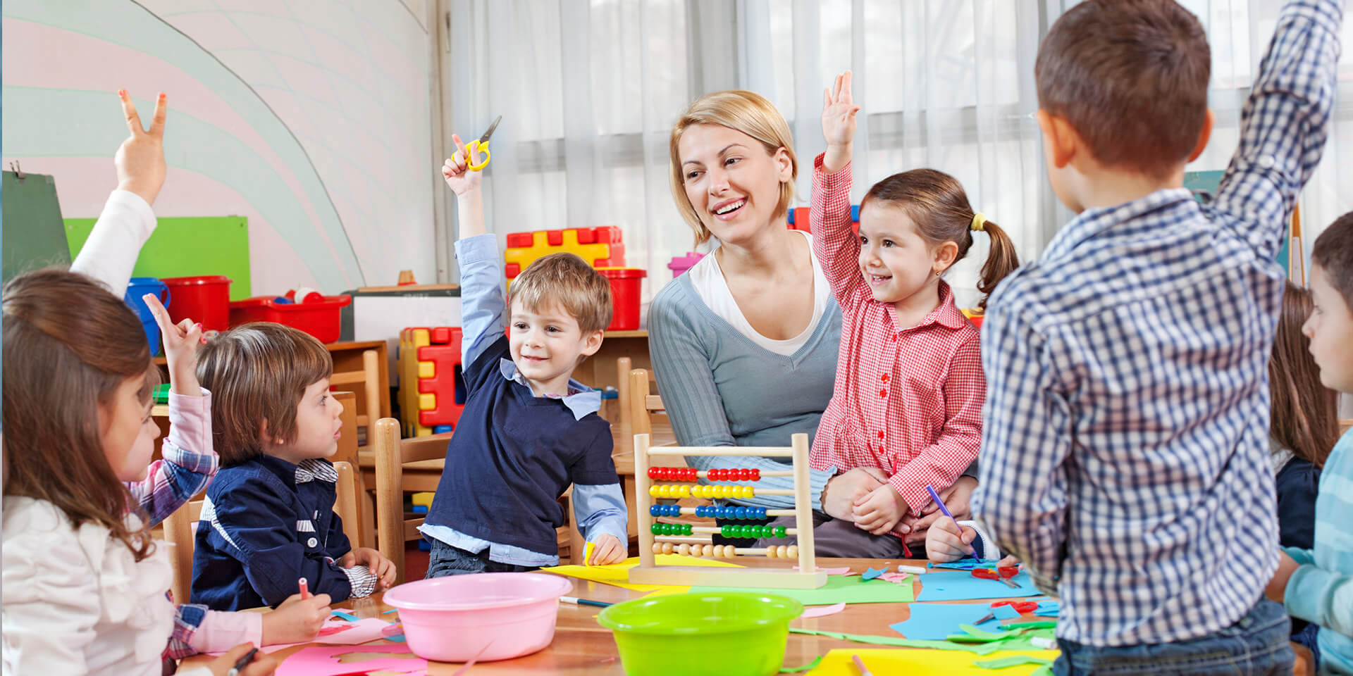 Das Bild zeigt eine Szene aus dem Kindergarten: eine Erzieherin sitzt mit Kindern um einen Tisch. Kindergarten