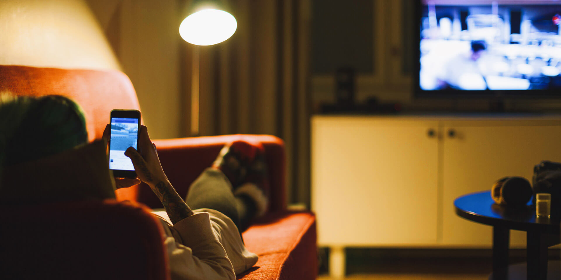 Person liegt auf dem Sofa und scrollt in dem Handy während der Fernsehen läuft Fernsehen mit zweiten Bildschirm
